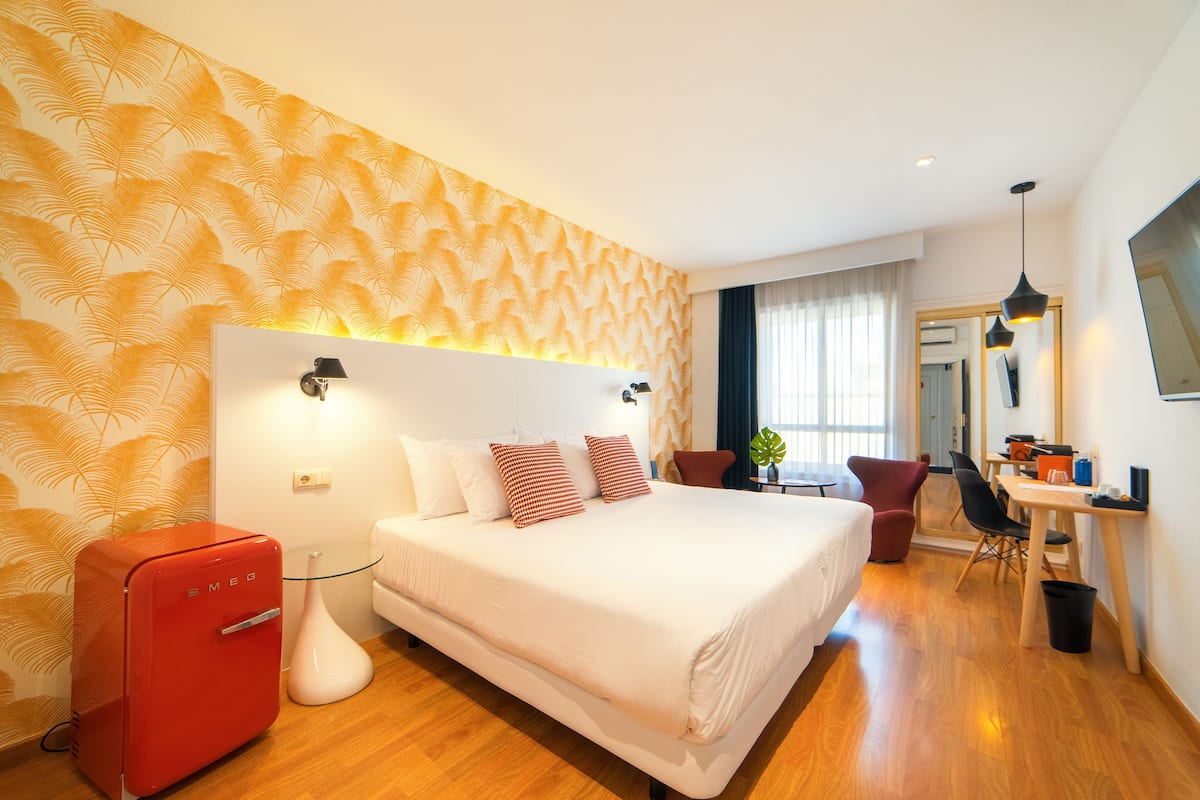 Une chambre d’hôtel moderne avec un grand lit, un bureau avec une chaise, un appareil électroménager rouge et un miroir au mur.