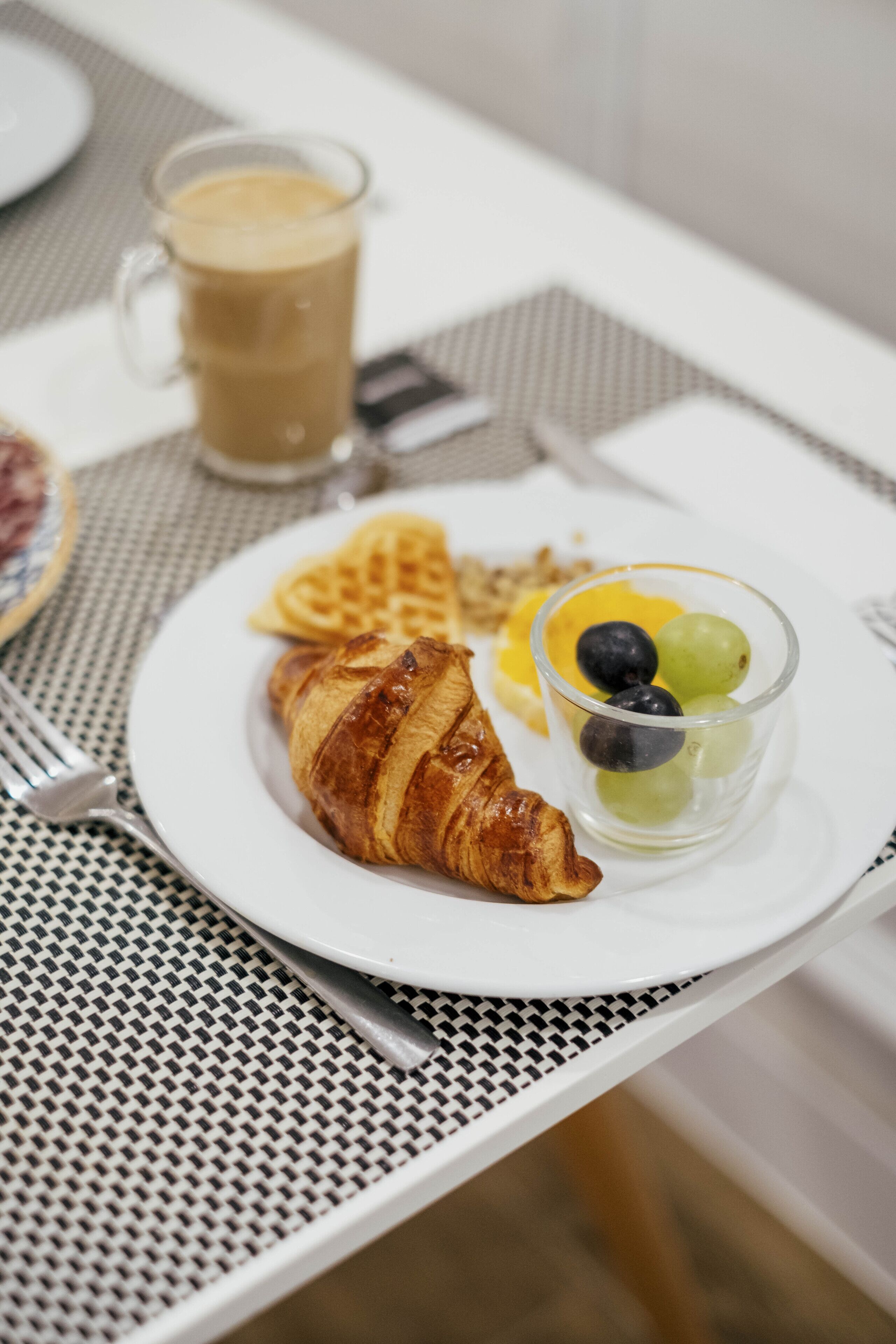 Un plato de desayuno con un croissant, gofres y un vaso de jugo de frutas sobre una mesa.