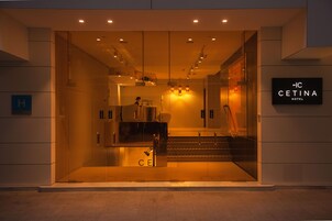 The entrance of the hotel with a glass facade, a reception desk, and a staircase leading inside.