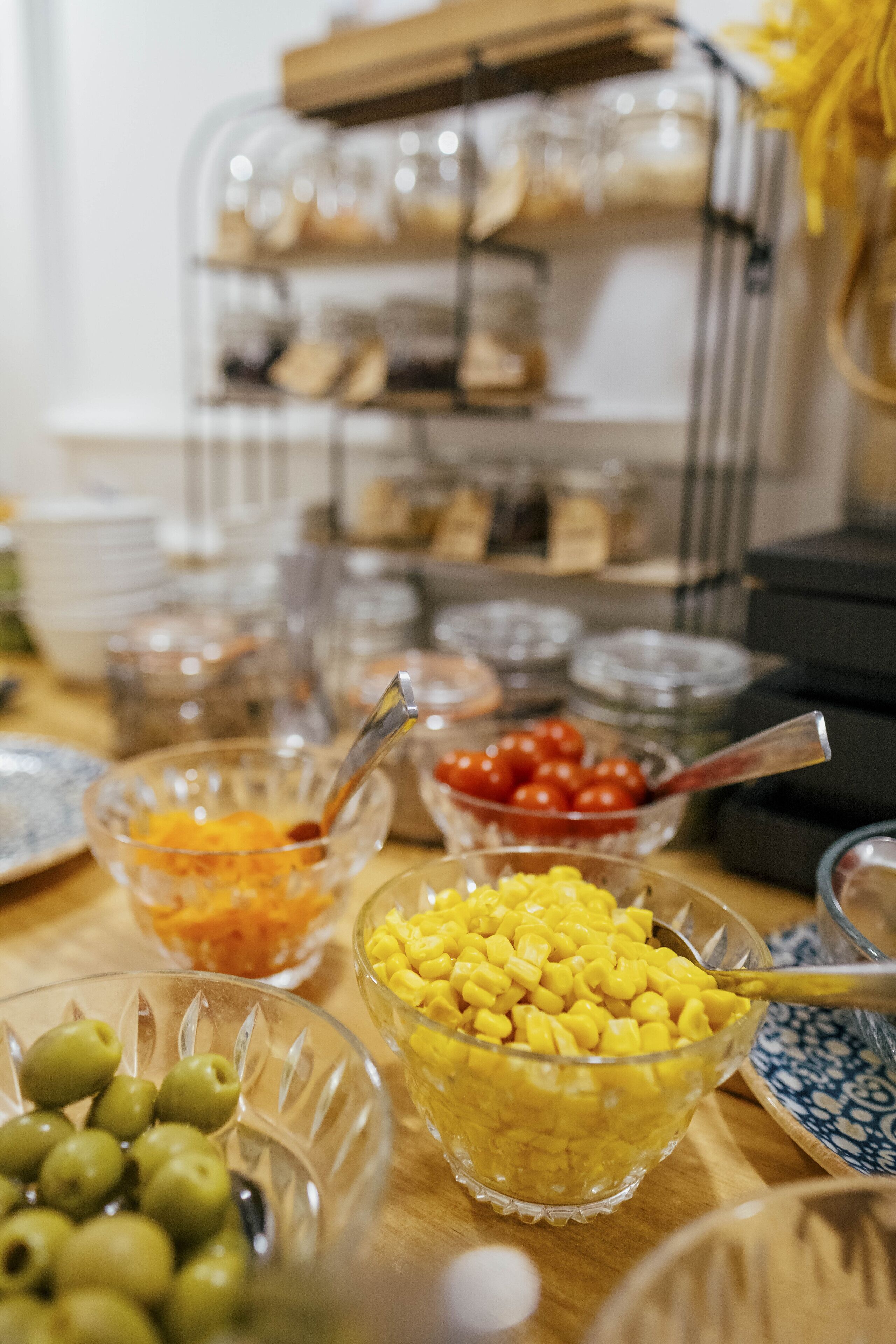 Una mesa de buffet con diversos alimentos en recipientes de vidrio transparente, como aceitunas, maíz y tomates cherry.