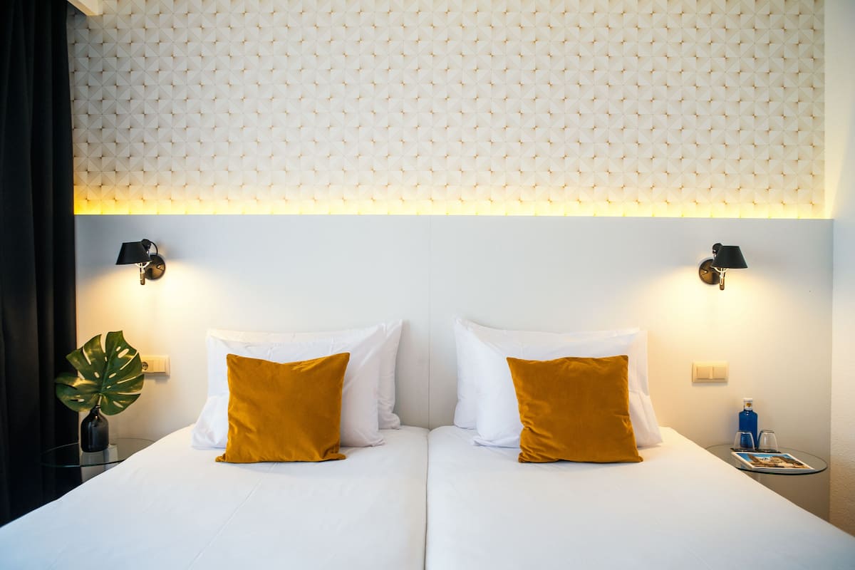 Une chambre d’hôtel avec un lit, deux oreillers, une table de chevet ornée d’une plante et des lampes fixées au mur.