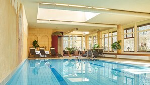 Une piscine intérieure à l’eau d’un bleu cristallin, baignée de lumière naturelle grâce à de grandes fenêtres, et entourée de chaises longues et de plantes en pot.
