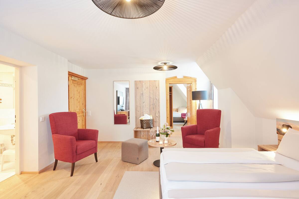 Ein modernes Hotelzimmer mit einem Bett, zwei roten Sesseln, einem kleinen Tisch mit einer Vase und einem Holzkabinett.