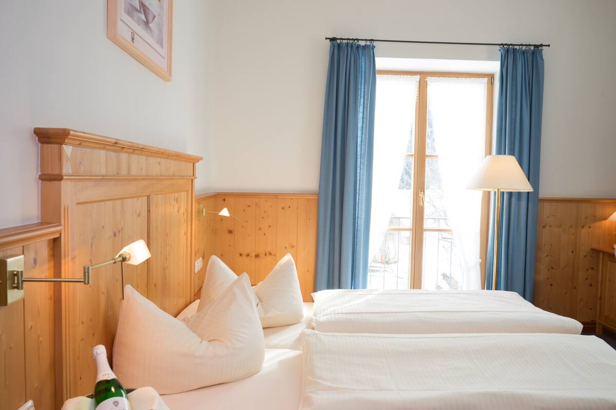 Ein Hotelzimmer mit zwei Betten, einem Holzkopfende, einem Nachttisch mit einer Flasche und einem Fenster mit blauen Vorhängen.