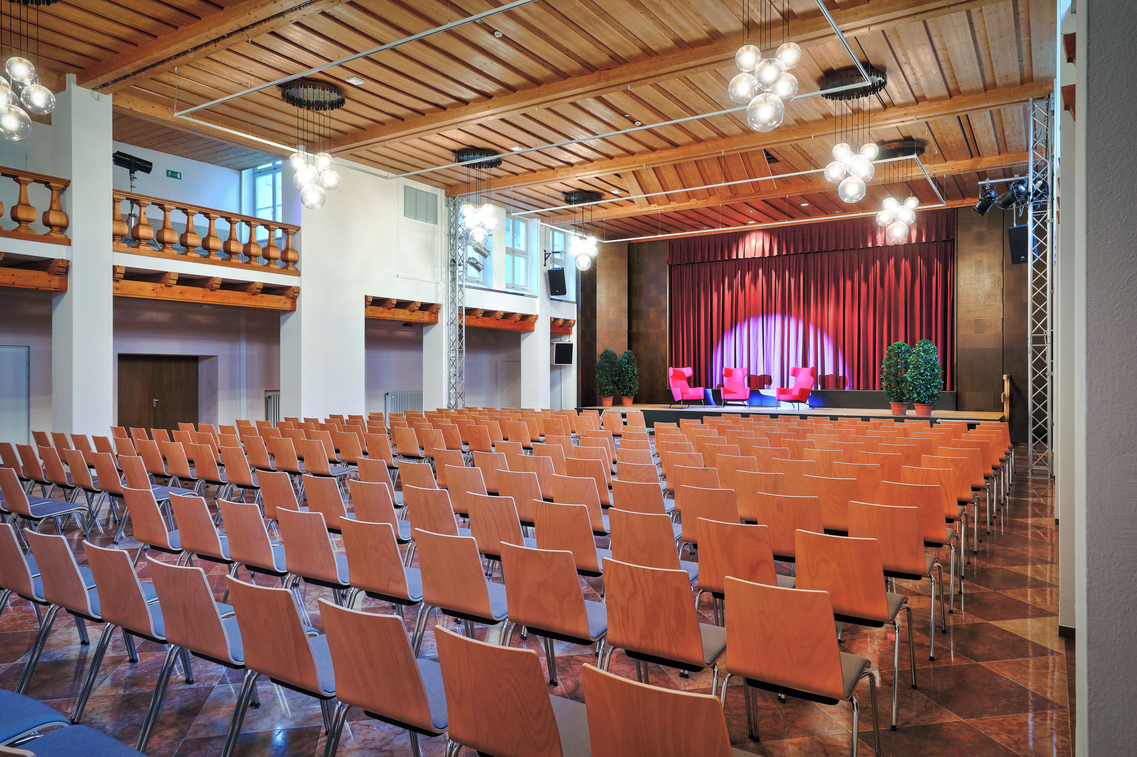 Een congrescentrum met rijen stoelen, een podium met gordijn en een balkon met houten leuningen.