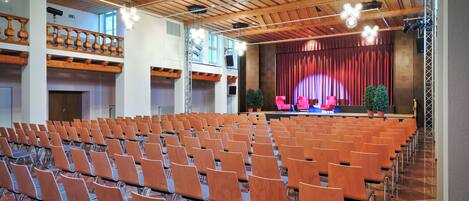 A conference room with rows of chairs, a stage with a curtain, and a balcony with wooden railings.