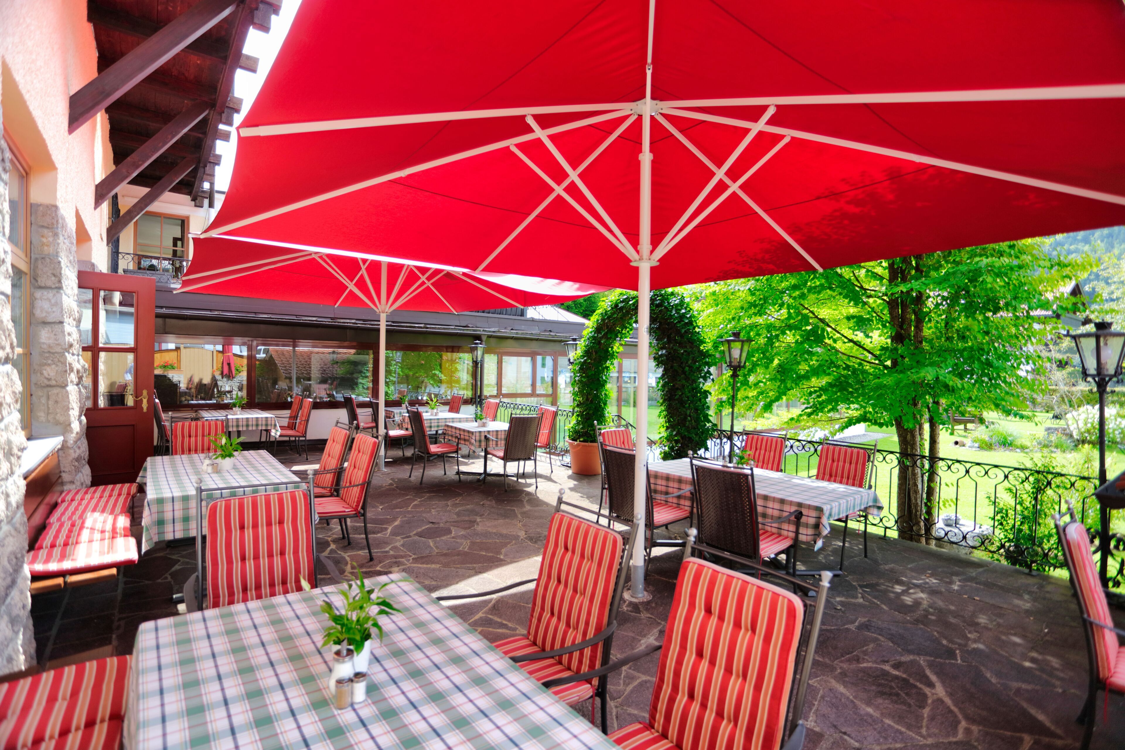 An outdoor patio area with tables and chairs, red umbrellas, and a view of greenery.