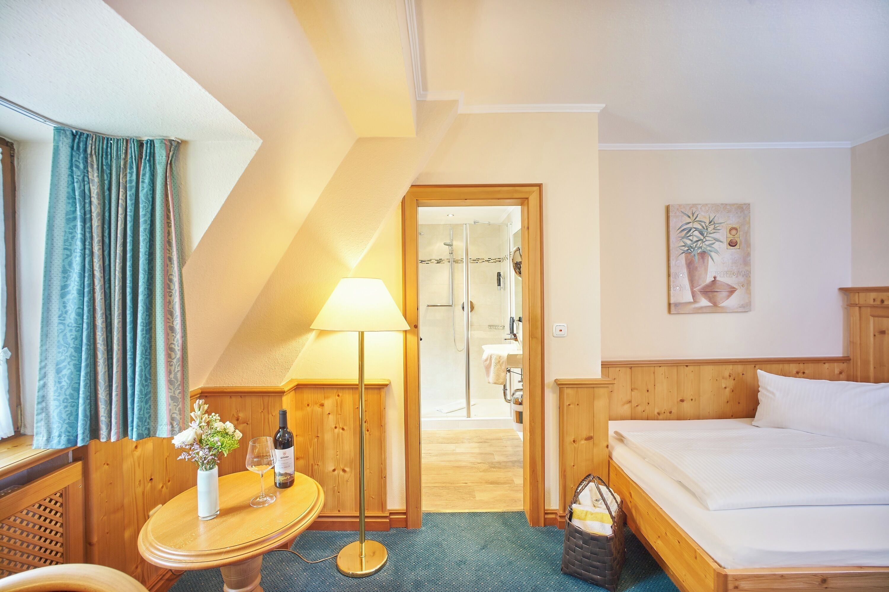 Une chambre d’hôtel avec un lit en bois, une petite table sur laquelle se trouvent un vase et une bouteille de vin, une lampe, et une vue sur la salle de bain et l’espace douche.