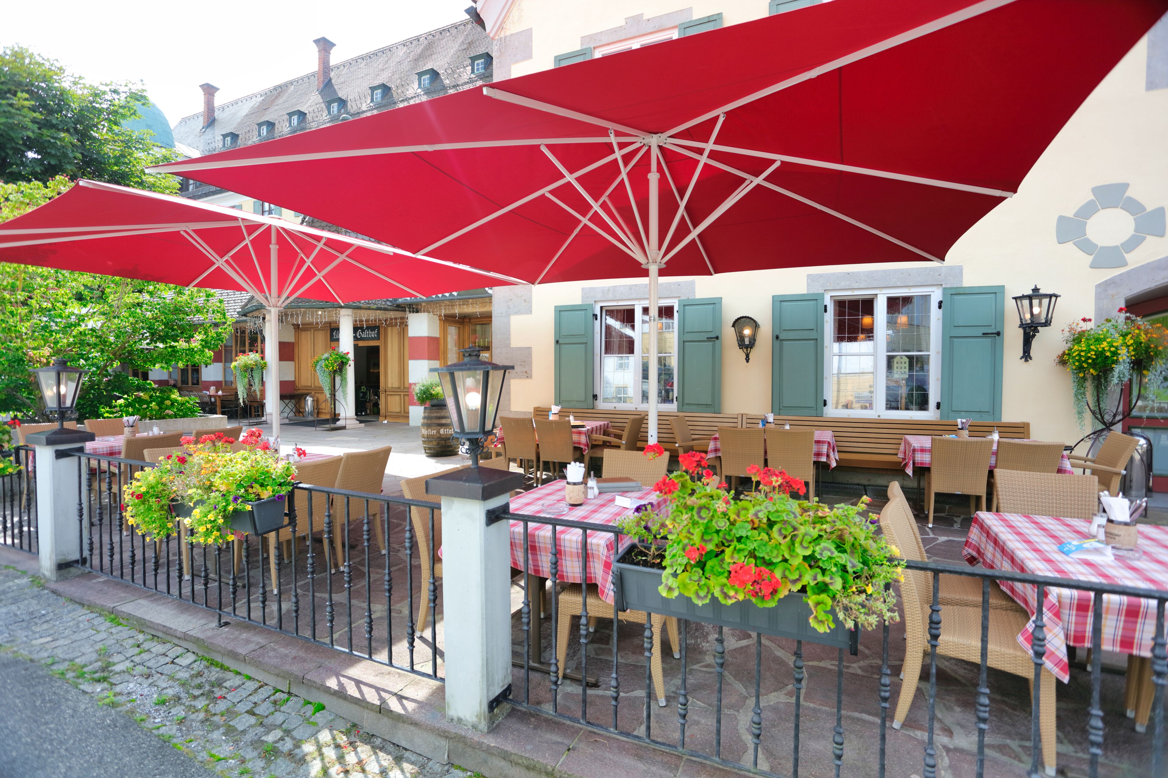 An outdoor cafe with red umbrellas, wicker chairs, and tables with checkered tablecloths.