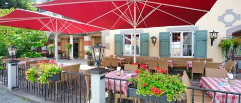 An outdoor cafe with red umbrellas, wicker chairs, and tables with checkered tablecloths.
