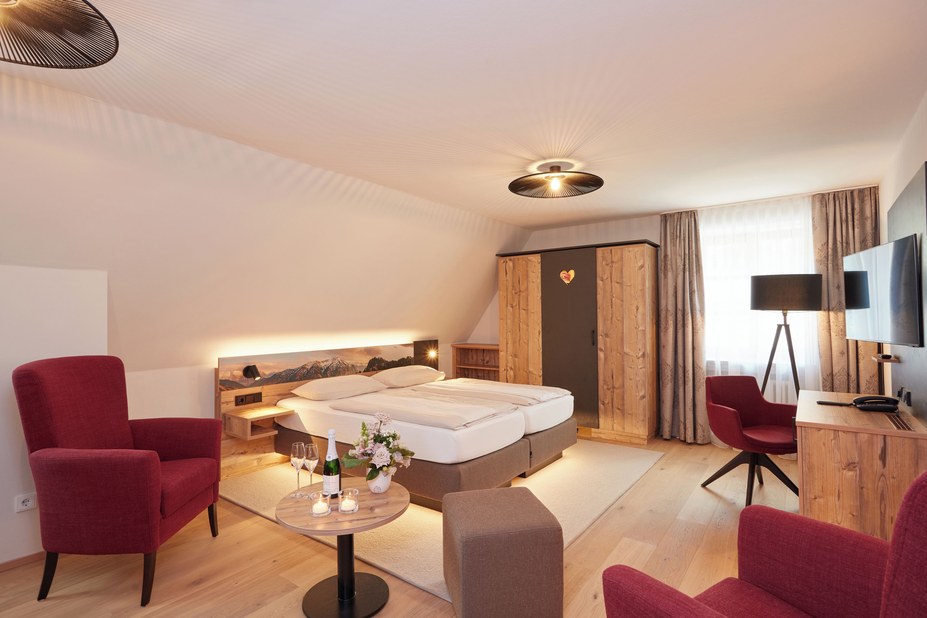 Una habitación de hotel moderna con una cama, dos sillones rojos, una mesita con un florero y velas, y un televisor sobre un mueble de madera.
