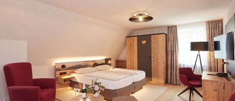Una habitación de hotel moderna con una cama, dos sillones rojos, una mesita con un florero y velas, y un televisor sobre un mueble de madera.