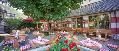 An outdoor seating area with tables and chairs, a flower bed with red flowers, and a building with a sign reading "LUDWIG DER BAYER".