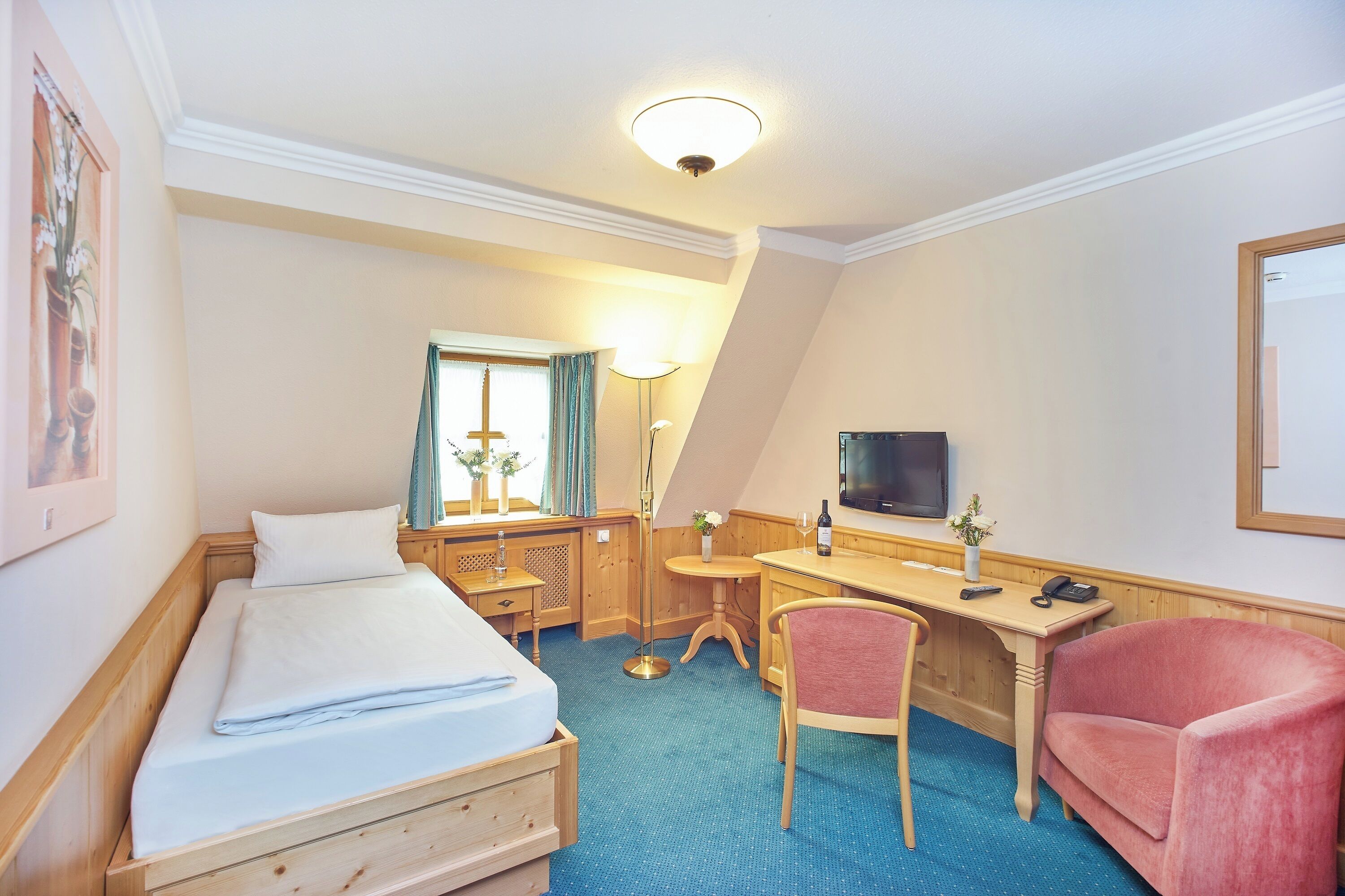 A hotel room with a bed, desk, chair, TV, and a small table with a vase and flowers.