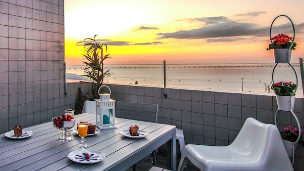 Une table à manger avec des assiettes remplies de nourriture et des boissons, une plante en pot, et une vue sur la plage au coucher du soleil.