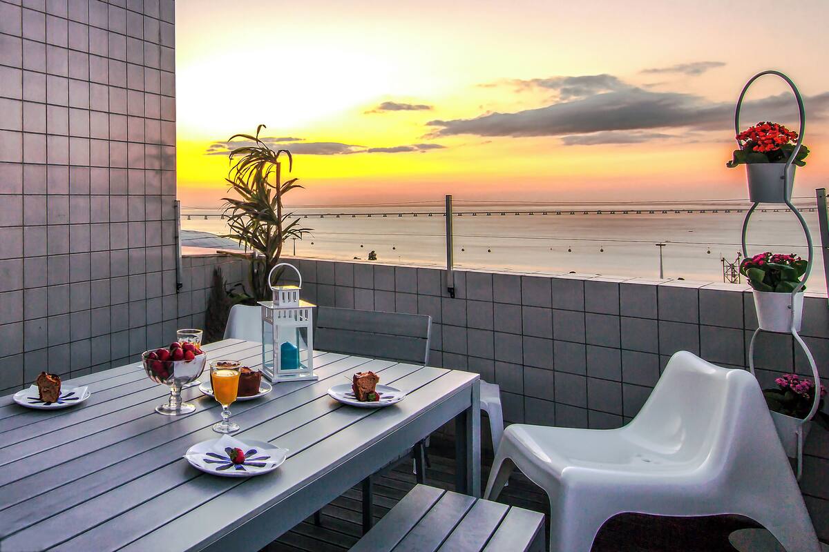 Una mesa de comedor con platos de comida y bebidas, una planta en maceta y vista a la playa al atardecer.