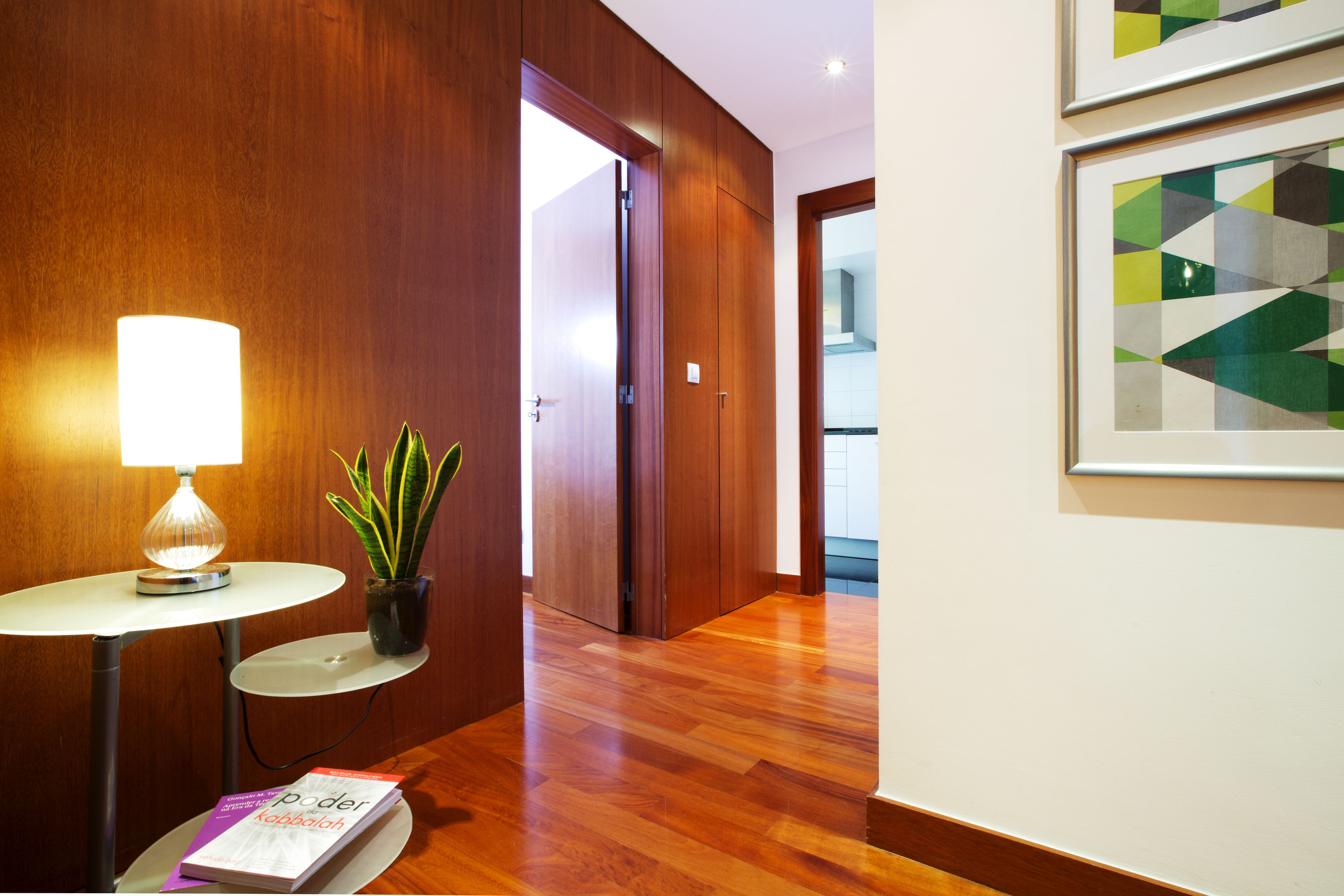 A hallway with wooden paneling, a round table with a lamp, a potted plant, and a framed artwork on the wall.