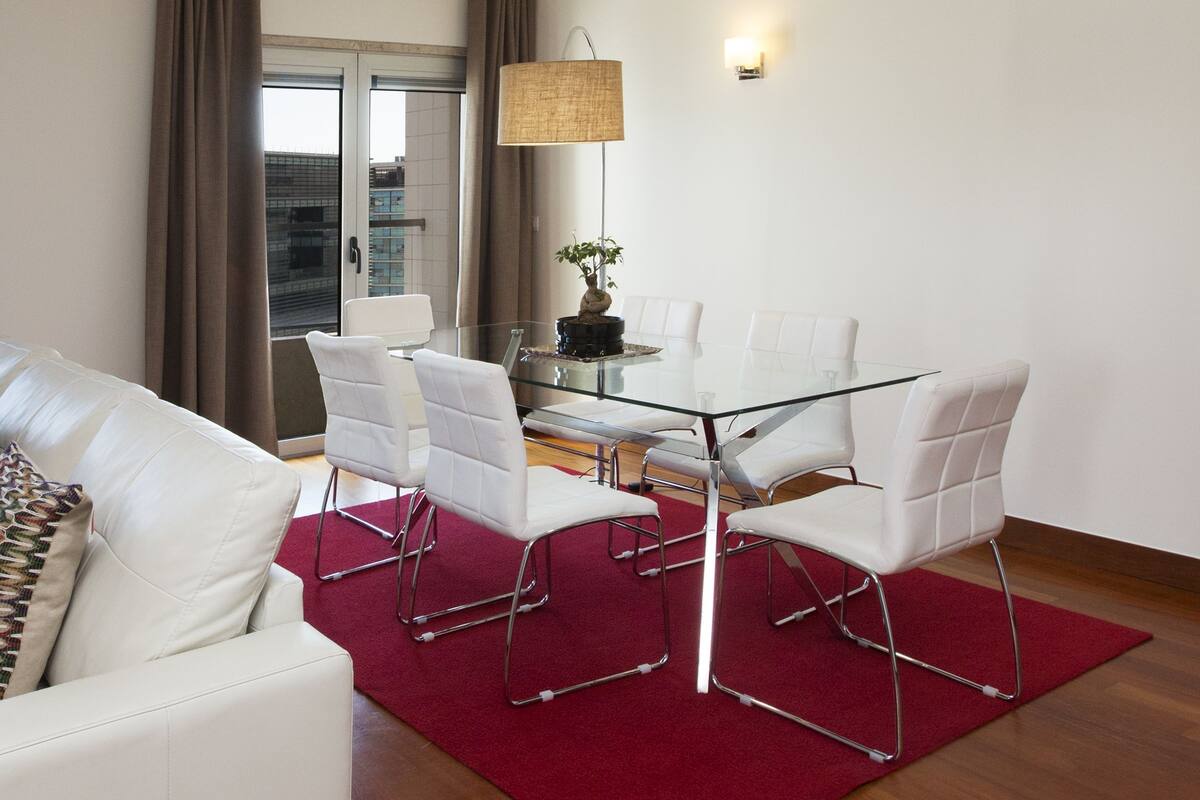 Una sala de estar moderna con una mesa de comedor de vidrio, sillas blancas, un tapete rojo y un gran ventanal con cortinas.
