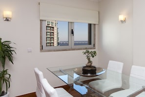 A modern dining area with a glass table, white chairs, a potted plant, and a view of a building across the street. - Oriente 25 Apartments by APT IIN - Parque das Nações (Lisbon)