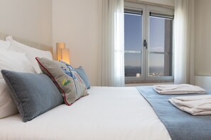 A hotel room with a bed, pillows, a window with a view, and a lamp.