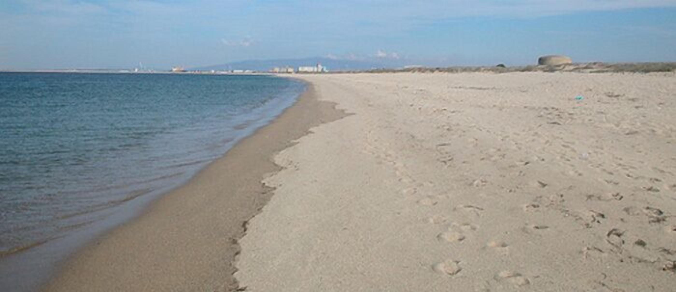 Een breed zandstrand met zachte golven, een heldere lucht en verre gebouwen.
