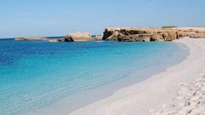 Una playa con agua turquesa cristalina, arena blanca y formaciones rocosas al fondo.