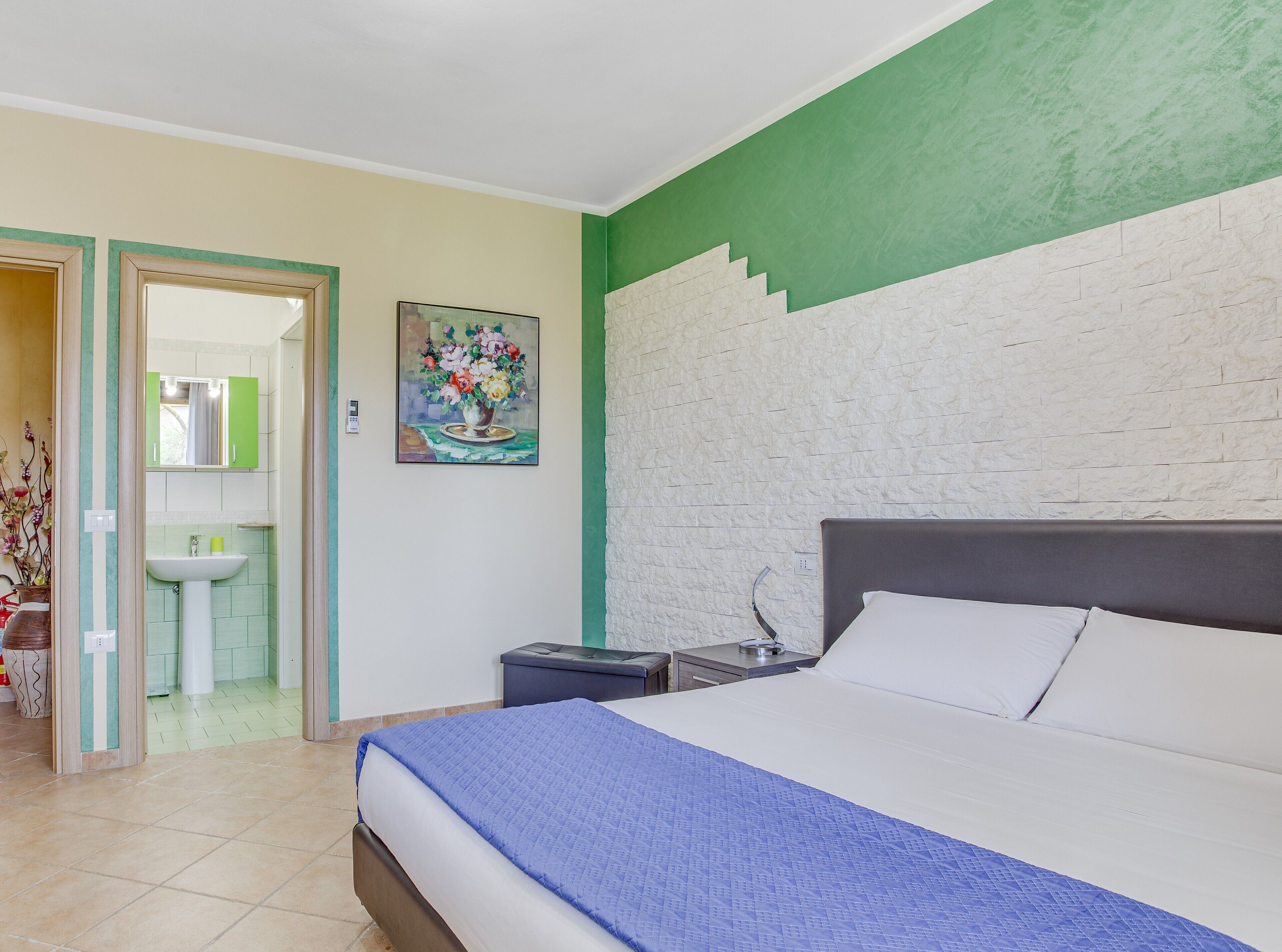 A bedroom with a bed, a nightstand, a painting, and a bathroom visible through an open door.