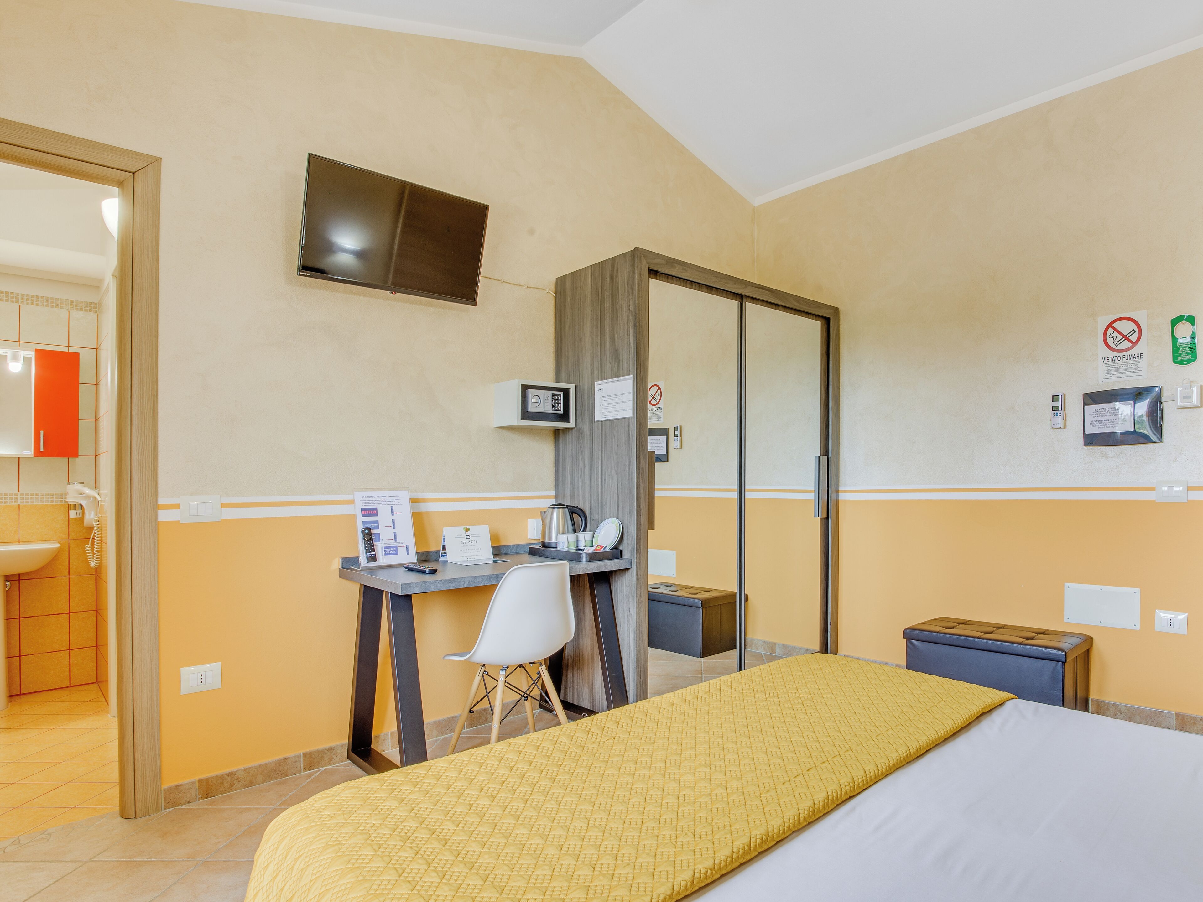 A hotel room with a bed, a desk with a chair, a television mounted on the wall, a mirror, and a small table with a kettle and cups.