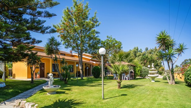Un jardín bien cuidado con una fuente, palmeras y un edificio amarillo con tejado de baldosas.