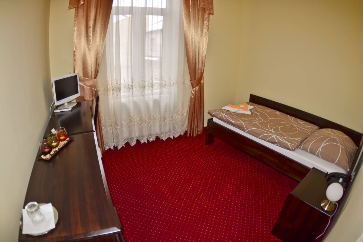 A hotel room with a wooden desk, a bed with patterned bedding, a red carpet, and sheer curtains.