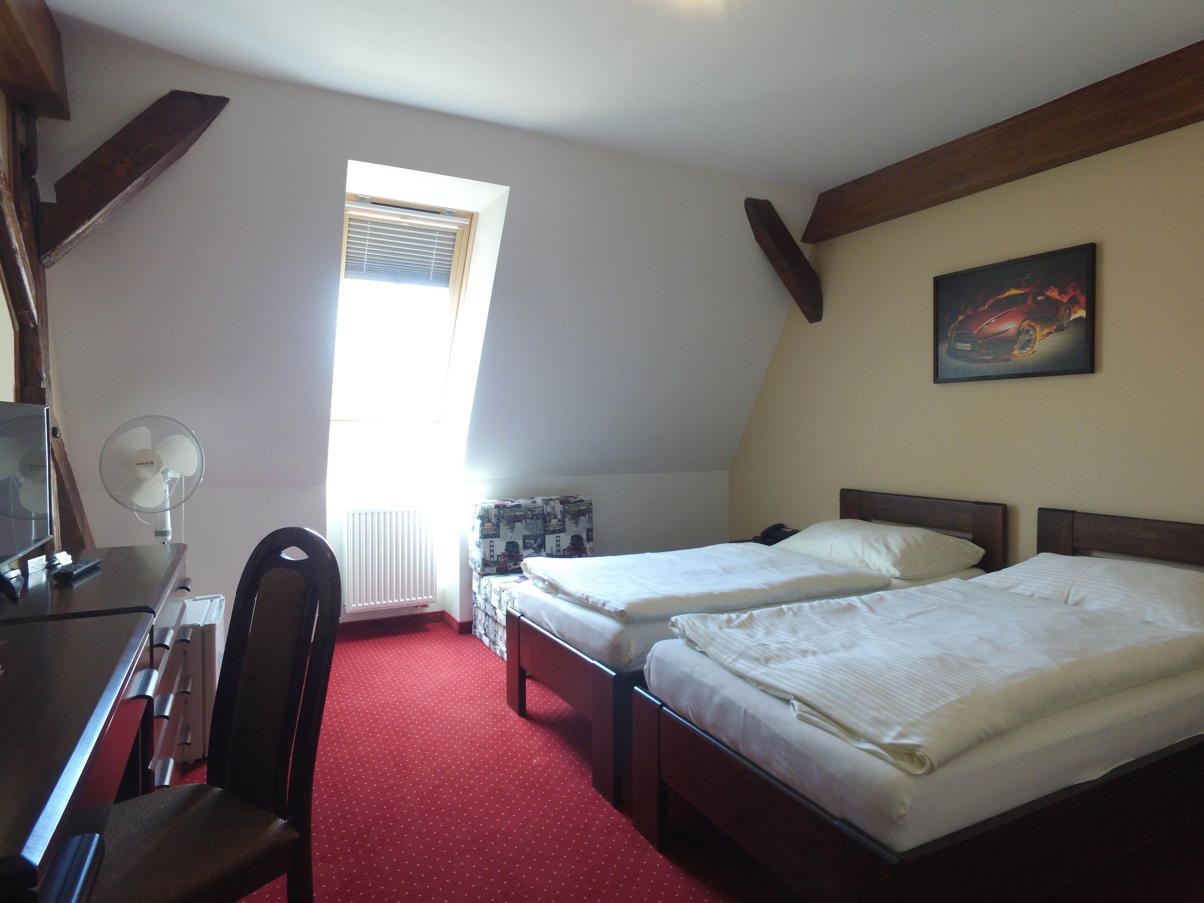A room with two beds, a desk with a computer and fan, a chair, a radiator, and a window with blinds.