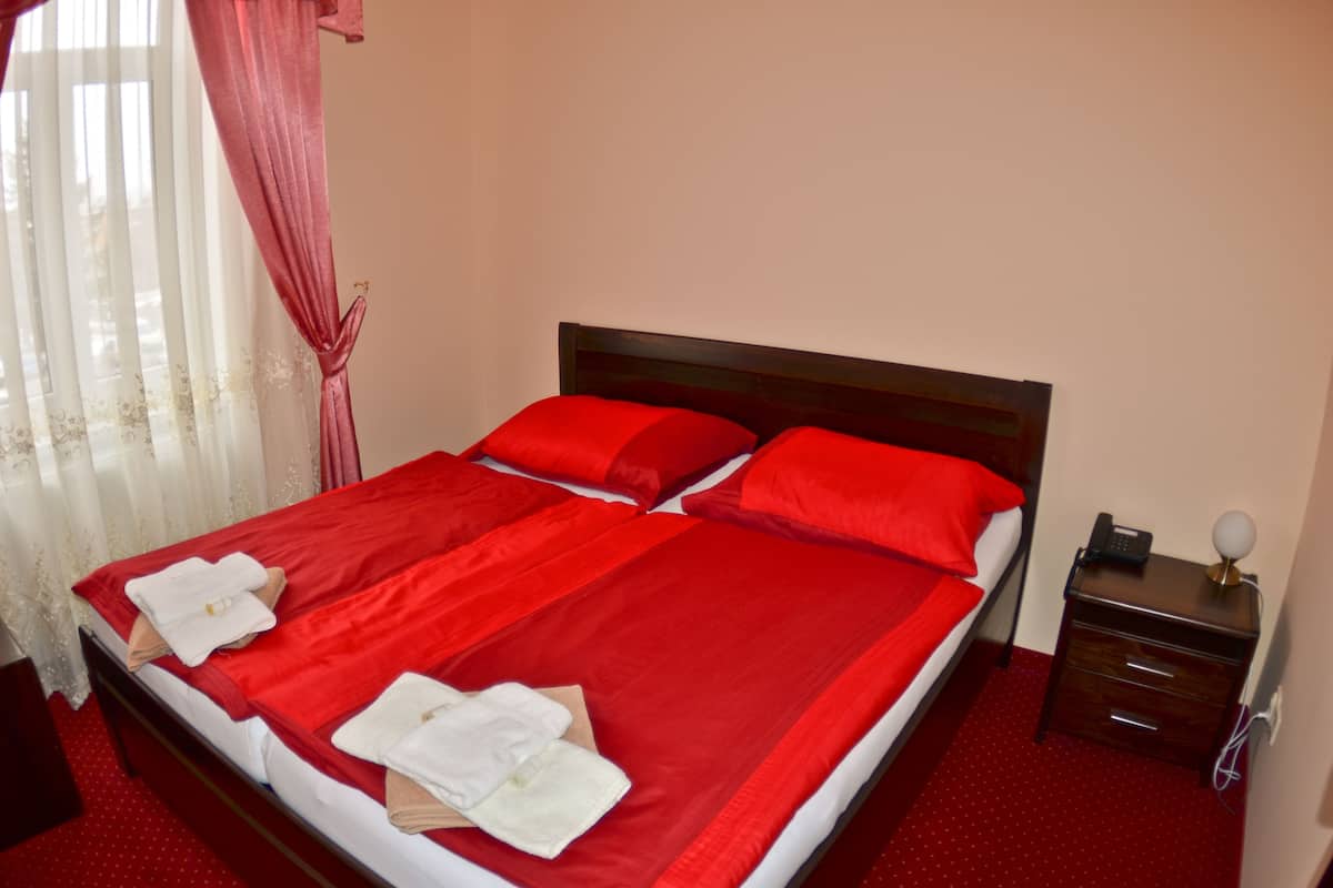 A neatly made bed with red bedding and white towels, a wooden nightstand with a lamp, and a window with sheer curtains.