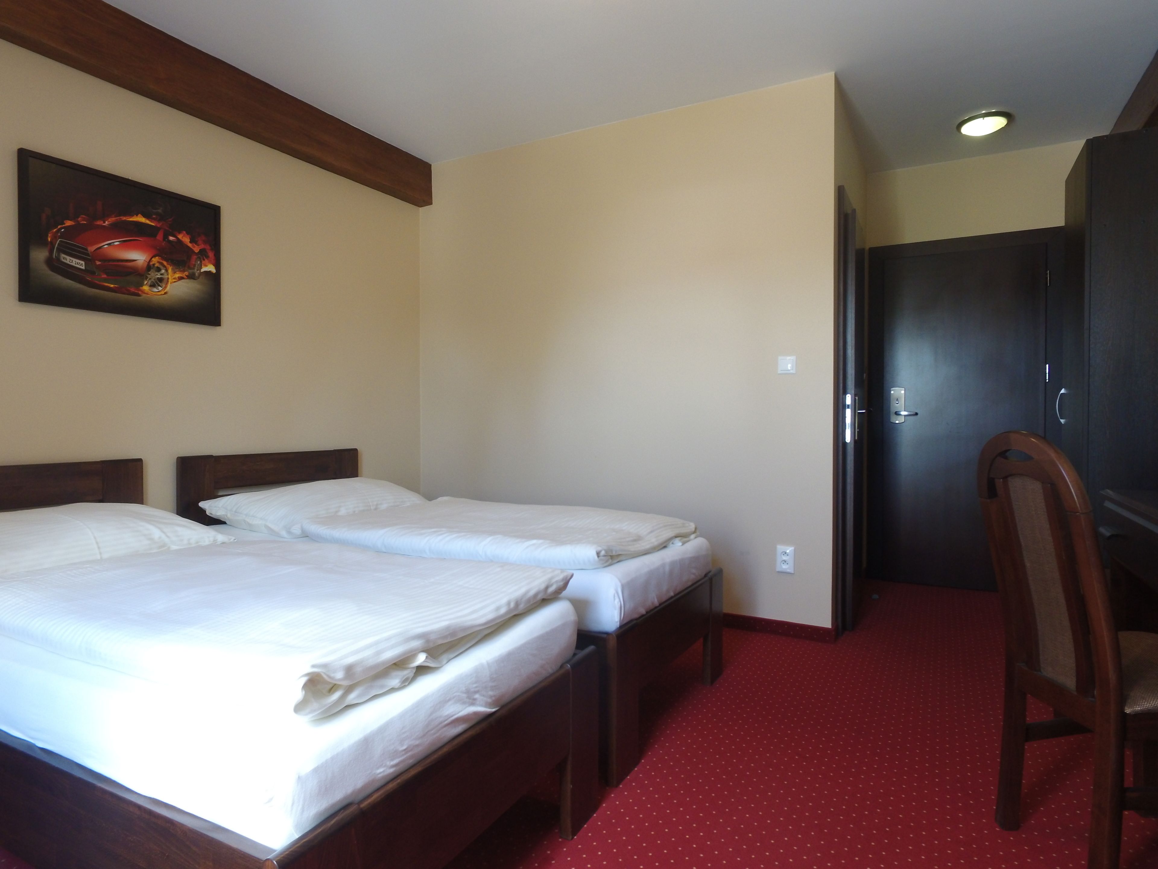 Une chambre d’hôtel avec deux lits simples, une tête de lit en bois, une moquette rouge, une porte et un tableau encadré au mur.
