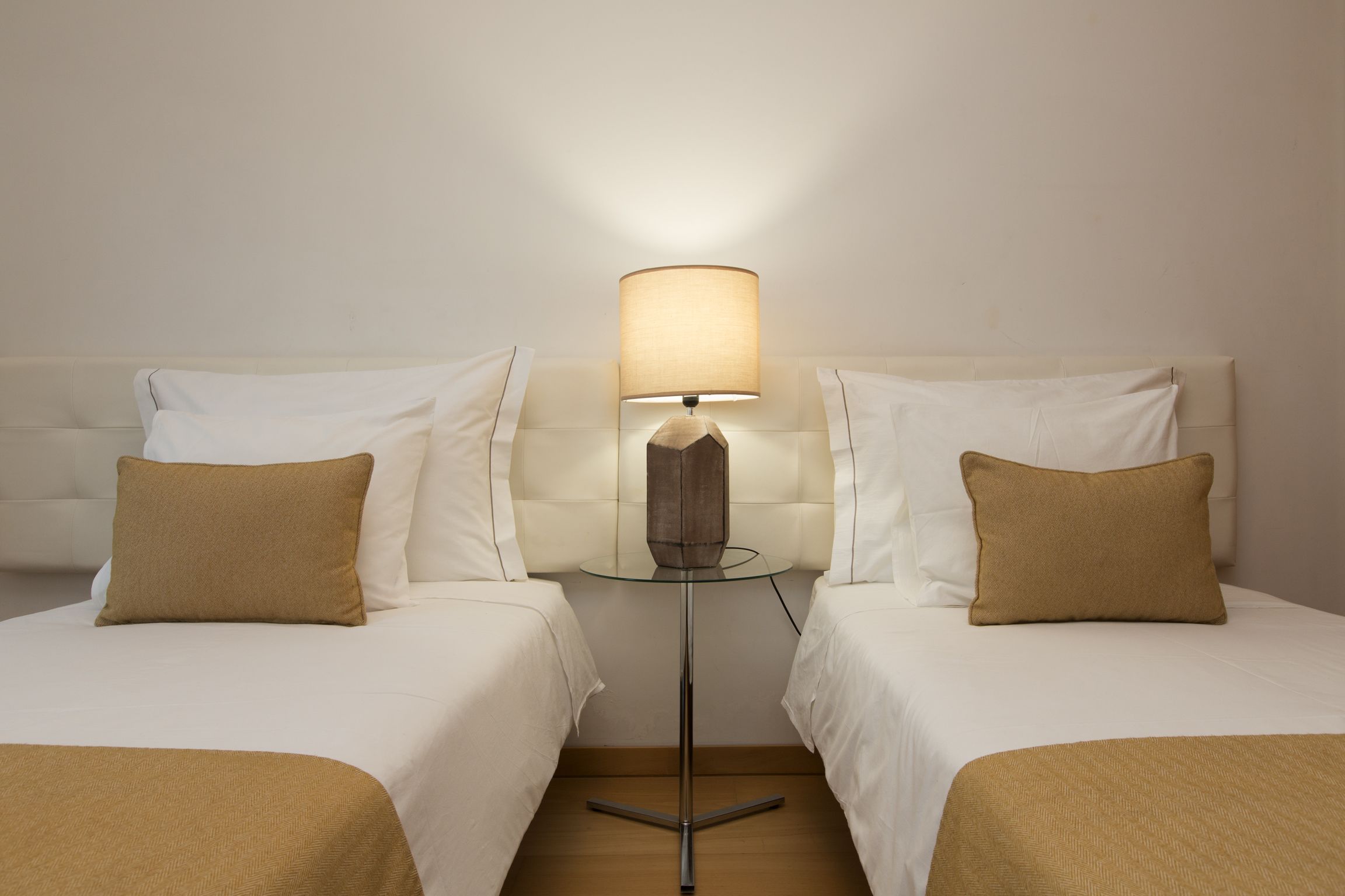 A hotel room with two beds, a small round table with a lamp, and a plain white wall.