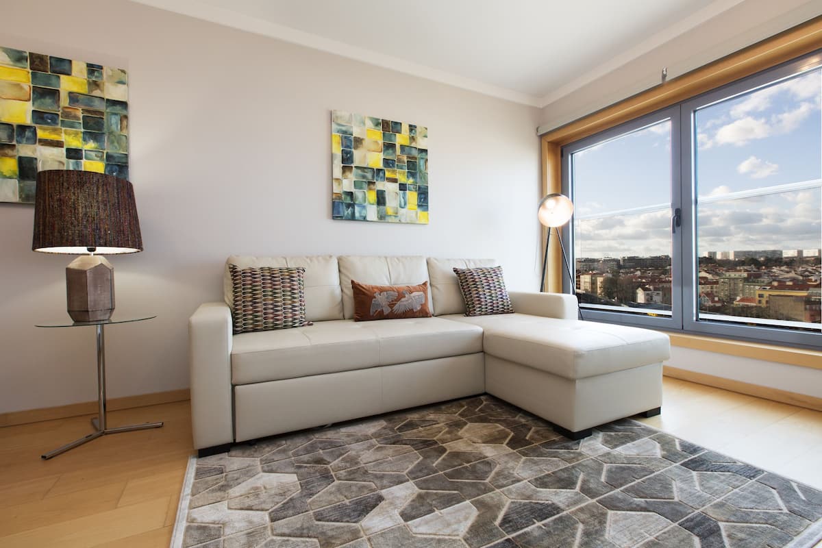 A modern living room with a sectional sofa, a patterned rug, and large windows offering a city view.