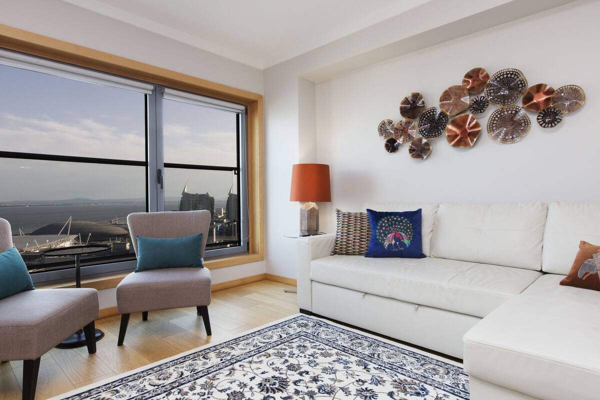 A modern living room with a white sofa, two armchairs, a patterned rug, and a large window with a view of a cityscape.