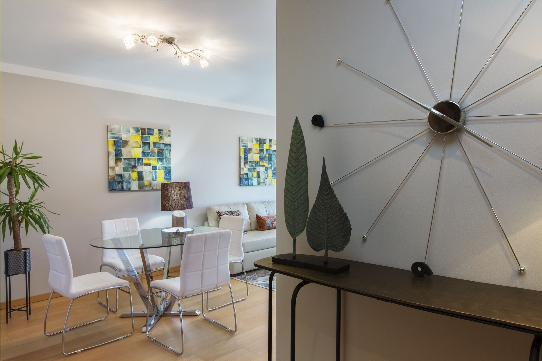 A modern dining area with a glass table and white chairs, a ceiling light fixture, and abstract art on the walls.