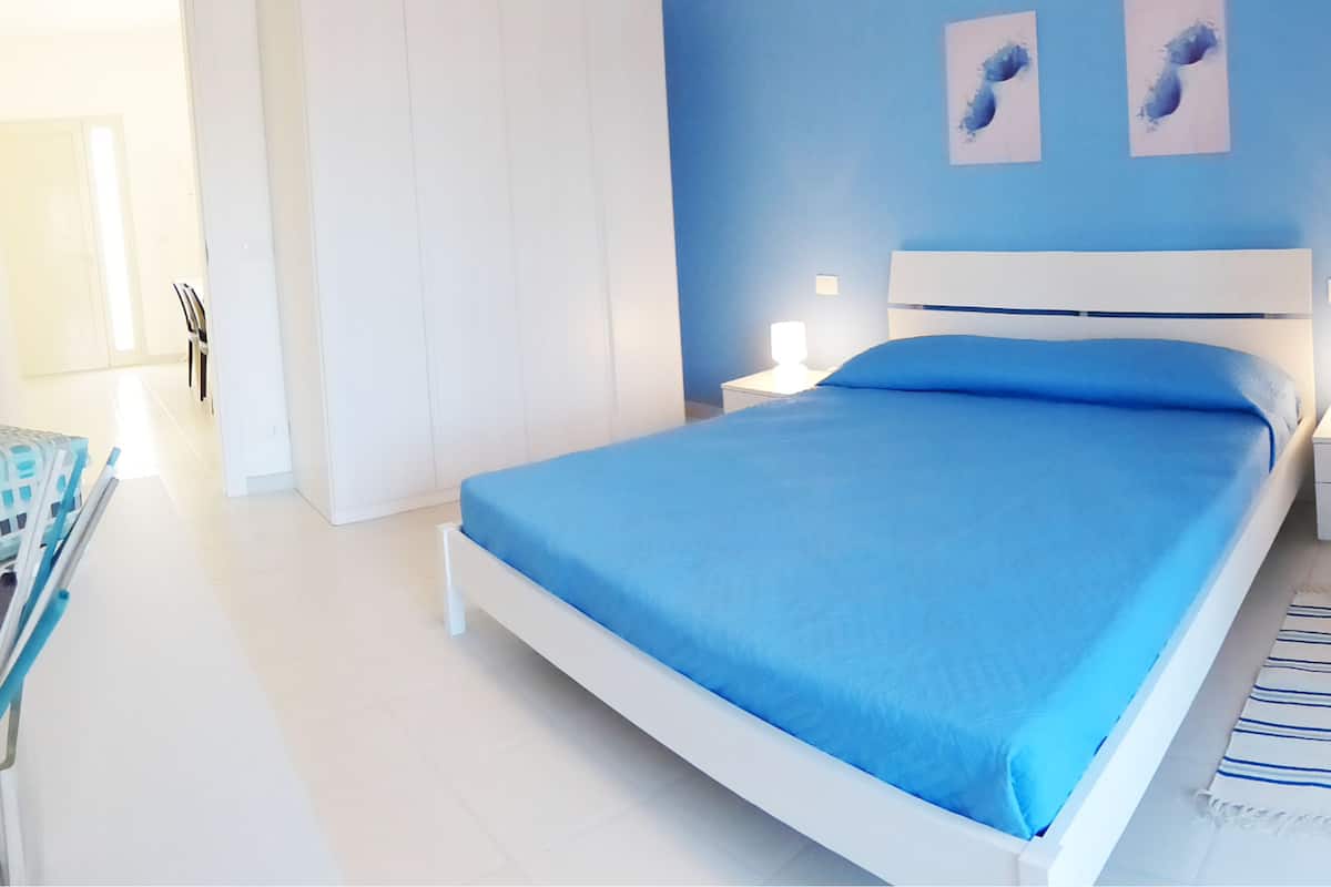 Un dormitorio con cama, mesitas de noche, una silla y cuadros en la pared.