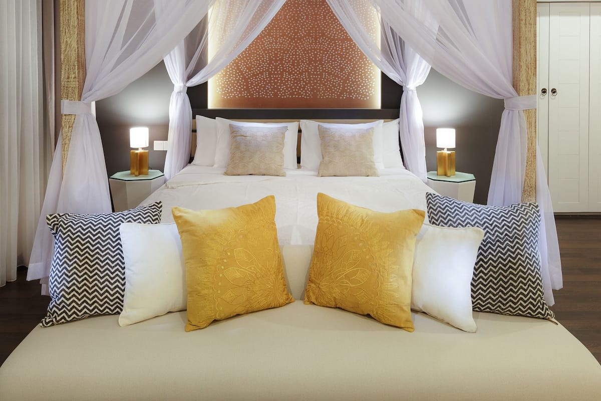 Uma cama grande com dossel, duas mesas de cabeceira com luminárias e almofadas estampadas.