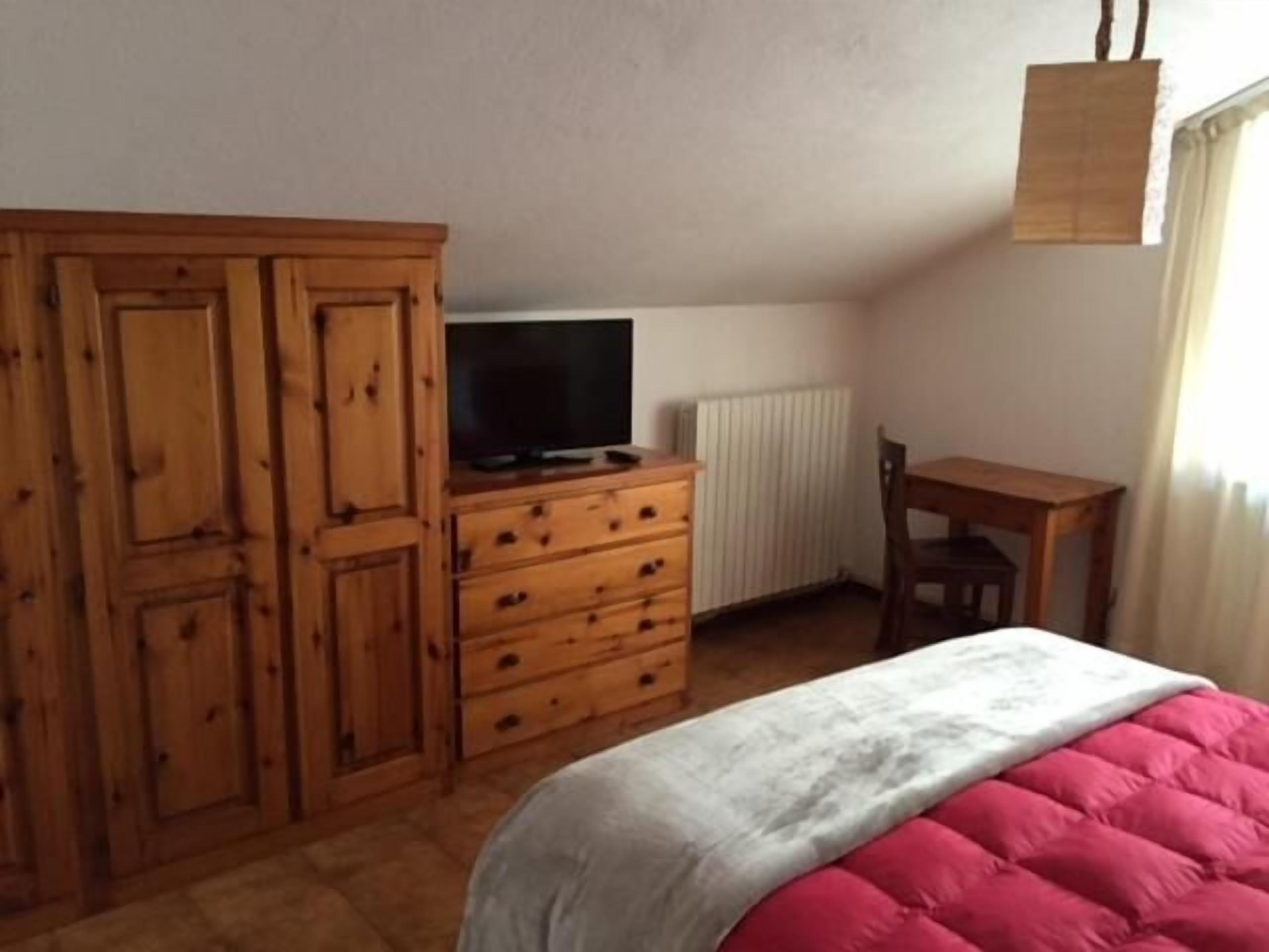 A bedroom with a bed, wooden wardrobe, a desk, and a hanging light fixture.