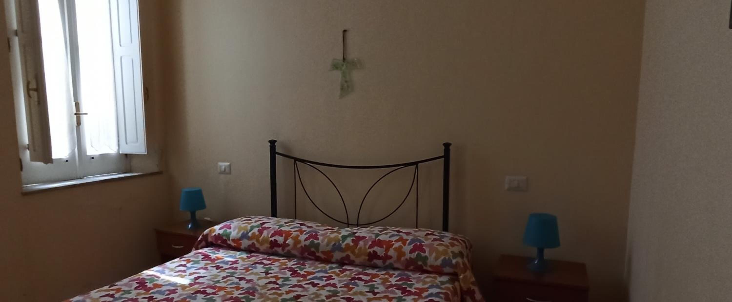 Een slaapkamer met een bed, twee nachtkastjes met lampen, een raam met gordijnen en een aan de muur bevestigd kruisbeeld.