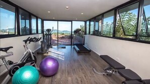 A modern gym with exercise equipment, large windows, and a view of the outdoors. - Dlux Condominium (Chalong)
