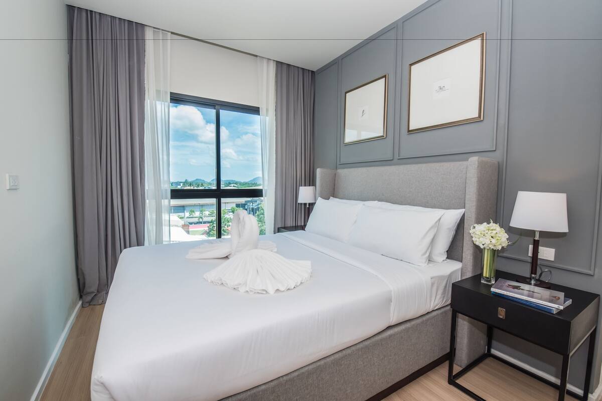 Una habitación de hotel con una cama grande, dos cuadros en la pared, una mesita de noche con una lámpara y vistas a la ciudad a través de la ventana.