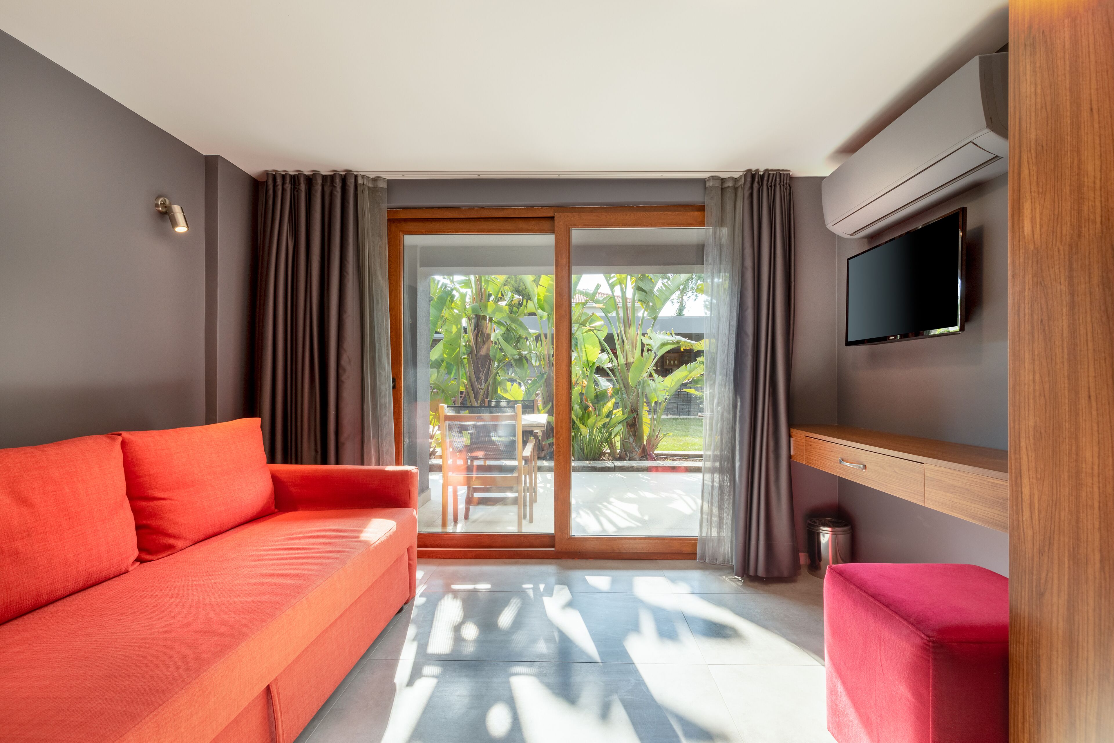 A modern hotel room with a red sofa, a small red ottoman, a flat-screen TV, and a sliding glass door leading to a balcony with plants.