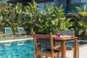 Una mesa de madera con cubierta de vidrio y una silla con cojín negro, ubicada junto a una piscina con vista despejada al agua.
