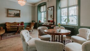 Una habitación con una mesa redonda de madera, varias sillas tapizadas, una araña, un cuadro y ventanas grandes con contraventanas.