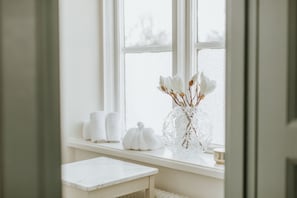 A bathroom with a white vanity, a window with white curtains, and a vase with white flowers on the windowsill. - Schenströmska Herrgården (Ramnas)