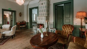 Una habitación con piso de madera, una mesa redonda con un florero, dos sillones, una araña, una chimenea y puertas verdes.