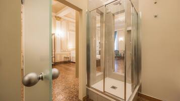 A hotel room with a glass-enclosed shower, a marble floor, and a view into the adjacent living area.