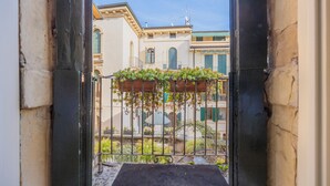 A view through a metal gate with a flower box, leading to a courtyard with a fountain and buildings in the background.